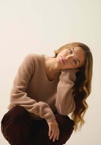 Beżowy sweter dziany z dekoltem w serek i długimi rękawami, połączony z ciemnobrązowymi spodniami sztruksowymi. Modelka pozując, opiera brodę na dłoni.