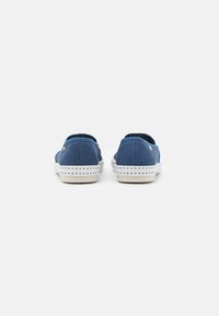 Blauwe canvas instappers met witte rubberen zolen, voorzien van stikdetails en een klein embleem aan de zijkant. Achteraanzicht toont een elastische opening.