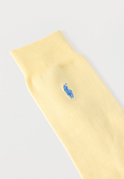 Ανοιχτόκίτρινη κάλτσα με μικρό μπλε κεντημένο λογότυπο αλόγου και αναβάτη κοντά στον αστράγαλο, εμφανίζεται σε λευκό φόντο.