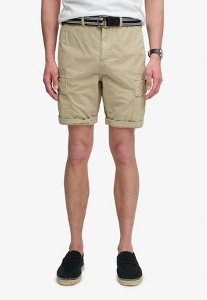 Man draagt beige opgerolde cargoshorts, gestreepte riem, wit T-shirt, zwarte slip-on schoenen en polshorloge, staande tegen een witte achtergrond.