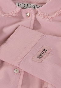 Roze overhemd met knopen en een kraag, met getaande kraagranden en een stoffen label op de mouw genaaid. Zachte textuur en een lichte tint.