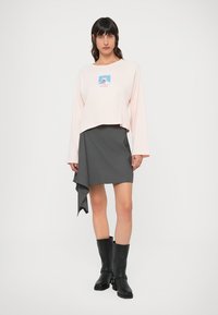 Lys rosa cropped langermet skjorte med et grafisk trykk, kombinert med en grå asymmetrisk omslagsskjørt. Sorte kne-høye støvler.