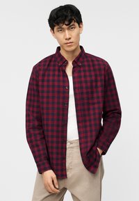 Νεαρός άνδρας που φοράει κόκκινο και μαύρο καρό πουκάμισο με κουμπιά πάνω από λευκή φανέλα, με μπεζ παντελόνι, στέκεται μπροστά σε μονόχρωμο φόντο.