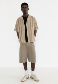 Lyse beige kortærmet skjorte med knapper og lodrette striber over en sort t-shirt, parret med beige shorts og cremefarvede sneakers med blå accenter.
