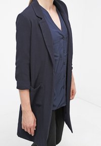 Marineblå lang blazer med skråklap, enkelt knaplukning, sidelommer og trekvartærmer, lavet af tekstureret stof.