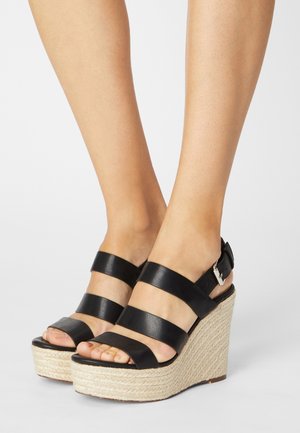 Des sandales compensées en cuir noir présentent trois larges brides avec une boucle ajustable, une plateforme tressée ressemblant à de la paille et une semelle texturée.