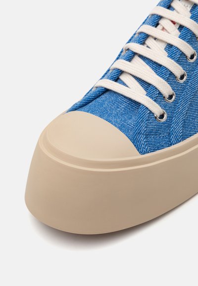Blue denim sneaker with a rounded beige rubber sole, featuring white laces and metal eyelets. Upper shows a textured fabric pattern.