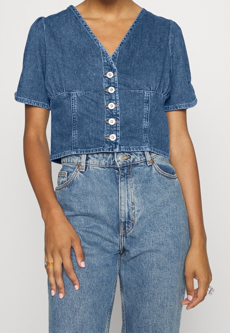 Femme portant un haut en denim bleu court à boutons avec des manches courtes bouffantes et un jean taille haute bleu clair.