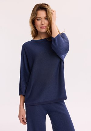 Kobieta z jasnobrązowymi, luźno falującymi włosami, ubrana w granatowy dzianinowy top i pasujące spodnie, delikatnie dotykająca włosów, na jednolitym tle.