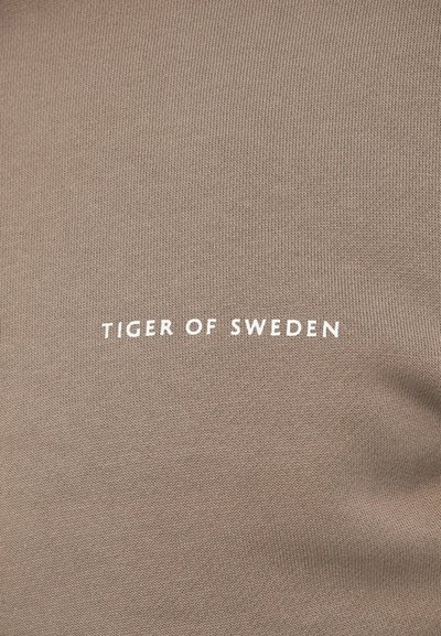 Beidzkrāsas audums ar baltiem "TIGER OF SWEDEN" burtiem, kas centrēti uz adījuma materiāla, parādot tekstūru un vieglas auduma krokas.