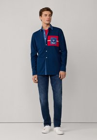 Camicia abbottonata di colore blu navy con una tasca sul petto rossa a contrasto, dettagli a righe bianche e colletto in denim; abbinata a jeans scuri e sneakers bianche.