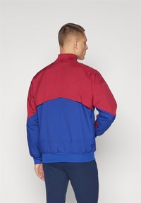 Fargeblokkert jakke med en rød øverste seksjon og en blå nederste seksjon. Laget av glatt stoff med ståkrage. Ribbede mansjetter.