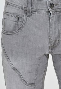 Jean en denim gris avec une coupe ajustée, style cinq poches et tissu texturé. Comprend une fermeture éclair et des détails de couture visibles.