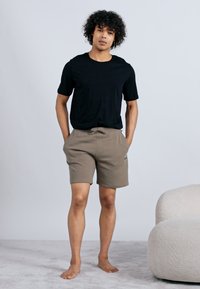 Tânăr cu păr creț, stând desculț, îmbrăcat cu un tricou negru și pantaloni scurți bej, pe un covor gri deschis, lângă un scaun alb cu textură.