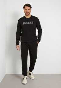 Felpa nera a maniche lunghe con il logo "GUESS" sul petto, abbinata a pantaloni neri e sneakers bianche con dettagli neri.