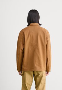 Giacca marrone con una vestibilità comoda, realizzata in tessuto resistente. Presenta un colletto, maniche lunghe e dettagli con bottoni vicino ai polsini. Vista posteriore visibile.
