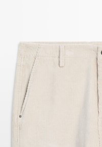 Pantalons en velours côtelé beige avec une texture à côtes verticales, dotés de poches latérales et d'une taille robuste avec une fermeture à bouton.