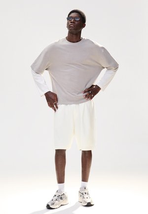 Mężczyzna w okularach przeciwsłonecznych, beżowa, luźna koszula z krótkim rękawem na białej, długiej koszulce, białe spodenki, białe skarpetki i białe trampki, stoi z rękami na biodrach.