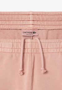Un pantalon de survêtement rose avec une taille à cordon, des détails côtelés et une étiquette Lacoste, mettant en valeur une texture de tissu douce et une coupe régulière.