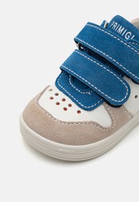Baskets blanches et beiges avec des bandes Velcro en suède bleu, des détails perforés et des coutures contrastées. Design basse avec une semelle en caoutchouc.
