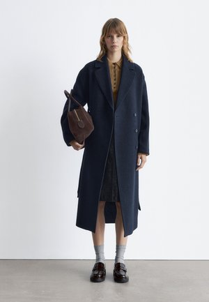 Navy wool blend overcoat with double-breasted design, wide collar, and side slits, paired with a brown leather bag and black loafers.