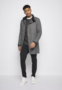 Abrigo gris texturizado con cuello negro, con cierre de cremallera, combinado con pantalones oscuros a rayas y zapatillas blancas.