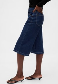 Mörkblå denimculottes med vida ben, med framfickor, hög midja och knappstängning, kombinerade med svarta slip-in-sandaler.