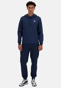 Felpa con cappuccio di colore blu navy, dotata di tasca a canguro e cordino, abbinata a pantaloni jogger coordinati e sneakers bianche con dettagli blu navy.