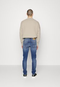 Beige sweatshirt med löst snitt, iklädd blå denimjeans med ljus tvätt och synliga sömmar. Svarta sneakers med vita accenter.