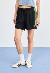 Svarte treningksorter med tre vertikale neon grønne striper, elastisk midje og brodert logo. Kombinert med hvite sokker og sko.