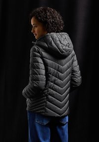 Svart puffjacka med zigzag-quiltad design, huva och dragkedjefickor, tillsammans med mörkblå jeans mot en svart bakgrund.