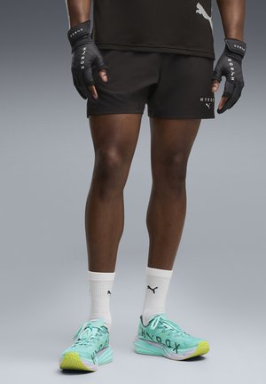 Jambes d'une personne portant un short noir HYROX, des gants noirs, des chaussettes blanches Puma et des chaussures de running HYROX turquoise avec des semelles jaunes.