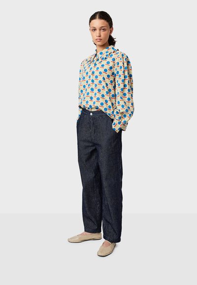 Blusa a maniche lunghe blu e arancione a pois, fit rilassato, con colletto. Pantaloni a righe scure, gamba ampia. Scarpe basse beige.