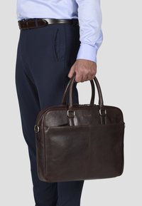 Brun lær laptopveske med doble håndtak, glatt tekstur, sølvfargede detaljer og en polert finish. Båret av en person i marineblå bukser.