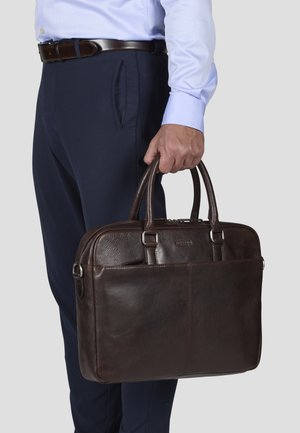 Brown leather laptop bag with dual handles, smooth texture, silver hardware accents, and a polished finish. Worn by a person in navy trousers.