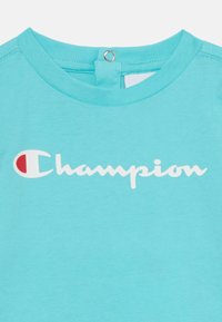 Heleda sinine puuvillane T-särk, millel on ümar kaelus ja nööbiga detail seljal. Ees on suur valge "Champion" logo.