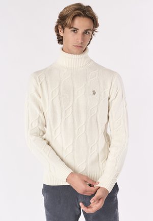 Giovane uomo con un maglione bianco a collo alto a coste e pantaloni scuri, che sistema la manica su uno sfondo chiaro e semplice.