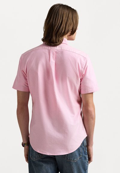 Chemise à manches courtes en tissu rose clair, avec un col classique, un pli central dans le dos et un ourlet arrondi, associée à un jean bleu.
