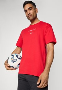 Rød bomulds t-shirt med rund halsudskæring og korte ærmer, med "ATLETI" trykt i hvidt på brystet. En mønstrede fodbold holdes i den ene hånd.