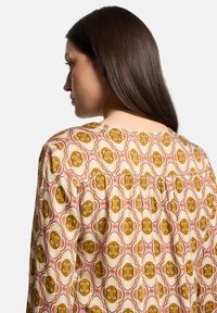 Blusa a maniche lunghe con un motivo floreale nei colori giallo, marrone e rosa. Presenta un retro arricciato e un colletto rotondo. Tessuto leggero.