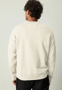 Sudadera de color crema con puños y dobladillo acanalados, que presenta un diseño sencillo en la parte trasera y una textura suave y apagada.