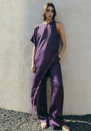 Donna che indossa un top asimmetrico senza maniche viola con rifiniture in pizzo e pantaloni a gamba larga abbinati, in piedi contro un muro beige strutturato.