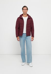 Mand iført en kastanjefarvet quiltet jakke, hvid t-shirt, lyseblå brede jeans og hvide sneakers, står foran en ensfarvet hvid baggrund.