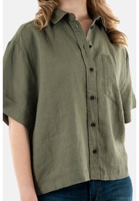 Camicia in lino verde oliva con maniche corte e chiusura a bottoni, dotata di tasca sul petto e vestibilità comoda con un orlo leggermente curvo.