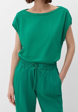 Žena nosící zelený top s krátkými rukávy zastrčený do vhodných kalhot s elastickým pasem a šňůrkou, stojí s jednou rukou v kapse.