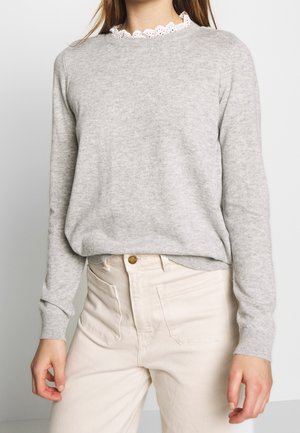 Femme portant un pull gris clair à manches longues sur une chemise blanche à col festonné en dentelle, associée à un pantalon taille haute écru.