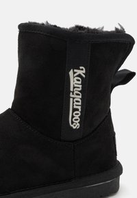 Černá jemná kožená kotníková bota s měkkou vnitřní podšívkou, vyznačující se výrazným bílým logem "Kangaroos" a poutkem pro snadné obouvání.