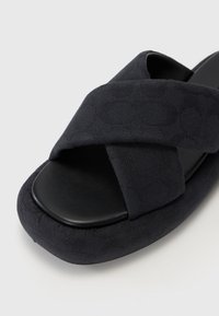 Svart sandaler med kryssremmer, laget av teksturert stoff på oversiden og glatt svart såle. Rund, polstret fotseng for komfort.