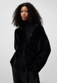 Zwarte faux fur jas met een relaxte pasvorm, voorzien van een brede kraag, knopen en een zachte textuur. Draagt over een zwarte coltrui.