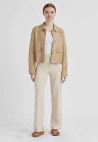 Cropped-Jacke aus braunem Leder mit großen Vorderaschen, weißem Tanktop, cremefarbenen, gerippten, weiten Hosen und braunen Rutsch-Sandalen.